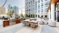 This contemporary amenity space is thoughtfully designed to blend upscale indoor comfort with resort-style outdoor living. The centerpiece of the interior is a soaring double-height living lounge wrapped in floor-to-ceiling glass curtain walls, flooding the space with abundant natural light and framing sweeping city skyline views. A dramatic stacked-stone fireplace anchors the room, complemented by warm wood-paneled ceilings, richly upholstered seating, and lush indoor greenery. A separate lounge area features slatted wood ceilings, soft neutral furnishings, a wall-mounted display screen, and organic sculptural wall art, creating an intimate yet refined setting. The interior opens seamlessly to a generous outdoor terrace finished in large-format stone pavers, where a fully equipped outdoor kitchen with professional-grade grills and a spacious dining table with string lighting invites al fresco entertaining. The expansive rooftop grounds include a curved, landscaped lawn edged by a sweeping concrete walkway, manicured garden beds, and mature trees — all set against a panoramic urban backdrop. A clean, gallery-style corridor with soaring ceilings and curated artwork connects the various areas, reinforcing the space's polished, modern aesthetic throughout.