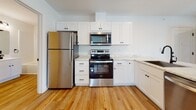 This single-floor unit offers a thoughtfully designed open-concept layout, where the living area and kitchen flow seamlessly together. Light hardwood flooring runs throughout the main living spaces, complementing the bright white walls and recessed lighting overhead. Large windows and a glass-paneled entry door allow generous natural light to fill the living area, creating an airy, welcoming atmosphere.

The kitchen is a standout feature, anchored by a peninsula with a shiplap-paneled base and a marble-look stone countertop that also incorporates a stainless steel undermount sink with a matte black faucet. White shaker-style cabinetry with matte black hardware lines the walls, paired with stainless steel appliances including a refrigerator, range, and over-the-range microwave. Ample upper and lower cabinet storage ensures functional workspace throughout.

The bedrooms are carpeted for comfort, each finished with crisp white walls. One bedroom features a set of double closet doors, providing generous storage. The bathroom presents a clean, modern aesthetic with white cabinetry, stone countertop, a tub-shower combination with white tile surround, and coordinating matte black fixtures throughout. A dedicated laundry area houses a full-sized top-loading washer and dryer with overhead shelving for added convenience.