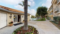 This residential complex presents a cohesive Spanish-inspired architectural design, characterized by warm stucco exteriors, clay tile rooflines, and arched entryways that establish a welcoming Mediterranean aesthetic. The community is organized around well-maintained common areas that connect multiple multi-story buildings, each accessible via exterior staircases and featuring private balconies with metal railings overlooking the grounds.

The landscaped courtyard serves as the heart of the community, featuring a lush green lawn, mature shade trees, tall palm trees, and manicured flower beds filled with colorful seasonal blooms. Wide concrete pathways wind throughout the property, linking residences to shared amenities. A dedicated leasing or management office sits conveniently near the entrance, identified by its inviting double doors and terra cotta tile roof.

A standout amenity is the generously sized outdoor swimming pool, enclosed by a secure fence and surrounded by a spacious pool deck furnished with shaded seating areas ideal for relaxation. Ground-level garage units line the rear of the property, providing covered parking. The overall ambiance is that of a well-kept, community-oriented environment with a distinct Southern California character.
