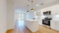 This well-appointed one-bedroom, one-bathroom unit offers a thoughtfully designed open-concept layout that maximizes every square foot. The combined kitchen and living area serves as the heart of the home, anchored by a sleek white kitchen island topped with a clean quartz-style countertop and illuminated by a pair of stylish gold pendant lights. The kitchen features crisp white cabinetry, open shelving, a white subway tile backsplash, and a full suite of stainless steel appliances including a refrigerator with ice maker, range, microwave, and dishwasher. Light hardwood flooring runs throughout the main living areas, adding warmth and continuity. The living room is bright and airy, equipped with a ceiling fan and French doors that open to a private balcony, allowing natural light to pour in and offering outdoor views. The bedroom is carpeted for comfort, includes a ceiling fan, and features a window with exterior views. It connects directly to a spacious walk-in closet with built-in shelving and hanging rods. The full bathroom presents a long single-sink vanity with white cabinetry, a tub-shower combo with tile surround, and matching light hardwood flooring, with convenient direct access to the walk-in closet.