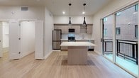 This single-floor residence showcases a thoughtfully designed open-concept layout with a cohesive, contemporary aesthetic throughout. Light hardwood flooring extends across all areas, complementing the neutral white and warm greige color palette that defines the space.

The kitchen serves as a central feature, equipped with stainless steel appliances including a refrigerator, gas range, and dishwasher. Light wood cabinetry with clean-line hardware pairs with white countertops, while open shelving adds a modern touch. A center island provides additional prep and seating space, and two bold black pendant lights hang above, adding a striking design contrast. A sliding glass door opens directly to a private balcony, bringing in natural light and outdoor views of the surrounding building architecture.

The bathroom is finished with the same light hardwood flooring and features a wood-tone vanity with white countertop, an illuminated mirror, and a full tub-shower combination. A separate bedroom offers a clean, neutral canvas with recessed lighting and ample wall space. A closet with louvered doors provides convenient storage. The overall ambiance is bright, modern, and move-in ready.