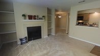 This single-floor home offers a comfortable and functional layout designed for everyday living. The living room serves as the heart of the space, anchored by a classic tile-surround fireplace with a white painted mantel — an ideal focal point that adds warmth and character to the room. Adjacent to the fireplace, a set of built-in corner shelves provides practical display and storage space, seamlessly integrated into the architecture. The room is finished with neutral wall-to-wall carpeting that extends throughout the living area, complemented by warm beige walls that create a cozy and welcoming atmosphere. A textured ceiling adds subtle dimension overhead, while clean white baseboards frame the space with a crisp finish. An open passageway leads further into the home, illuminated by a hanging pendant light that hints at a dining or transitional area beyond. A pass-through window visible on one side suggests a connection to an adjacent kitchen, enhancing the open, communicative flow between spaces. The overall ambiance is calm and neutral, offering a versatile backdrop well-suited for a variety of interior styles and personal touches.