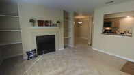 This single-floor home offers a comfortable and functional layout designed for everyday living. The living room serves as the heart of the space, anchored by a classic tile-surround fireplace with a white painted mantel — an ideal focal point that adds warmth and character to the room. Adjacent to the fireplace, a set of built-in corner shelves provides practical display and storage space, seamlessly integrated into the architecture. The room is finished with neutral wall-to-wall carpeting that extends throughout the living area, complemented by warm beige walls that create a cozy and welcoming atmosphere. A textured ceiling adds subtle dimension overhead, while clean white baseboards frame the space with a crisp finish. An open passageway leads further into the home, illuminated by a hanging pendant light that hints at a dining or transitional area beyond. A pass-through window visible on one side suggests a connection to an adjacent kitchen, enhancing the open, communicative flow between spaces. The overall ambiance is calm and neutral, offering a versatile backdrop well-suited for a variety of interior styles and personal touches.