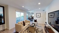 This contemporary single-floor residence offers a thoughtfully designed open-concept layout that seamlessly connects the main living areas. The living room serves as the heart of the space, featuring recessed lighting set into clean white ceilings, light wood-toned flooring, and a large sliding glass door that invites abundant natural light and provides direct access to an outdoor balcony area with greenery views. The living area is appointed with a neutral-toned sofa, a sleek glass-top coffee table, and a bold marbled area rug that anchors the space with a touch of sophistication. A framed artwork piece adds a curated, gallery-like quality to the walls. The open layout flows naturally into a dining area and kitchen beyond, where pendant lighting highlights a modern kitchen with dark cabinetry and a dining set that completes the entertaining space. The consistent color palette of whites, grays, and warm neutrals throughout creates a cohesive and inviting ambiance. Recessed lighting throughout ensures the space is well-lit at all times. Overall, this residence combines modern design sensibility with practical functionality, offering a comfortable and visually refined living environment.
