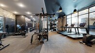 Located at 2010 N Brevard St in Charlotte's NoDa neighborhood, this contemporary fitness center is thoughtfully designed to accommodate a wide range of workout disciplines. The space features a striking interior with dark teal ceilings, warm pendant lighting, exposed stone accent columns, and a combination of light wood-tone and carpet flooring that creates a sleek, modern ambiance.

The cardio zone is positioned along a floor-to-ceiling glass window wall, providing ample natural light and outdoor views while housing a row of treadmills, rowing machines, and stationary bikes. A mounted flat-screen television and a water station offer added convenience during workouts.

The strength training area is exceptionally well-equipped, featuring Hammer Strength power racks, a multi-cable functional trainer, adjustable benches, and an extensive dumbbell rack. Additional functional training tools include medicine balls, battle ropes, and a heavy punching bag. Mirrored walls throughout enhance the sense of space and support proper form during exercise.

The layout flows intuitively, with clearly defined zones for free weights, machine-based strength training, and cardiovascular conditioning. The overall design balances industrial and modern aesthetics, making this fitness center a well-appointed and inviting space for a comprehensive training experience.