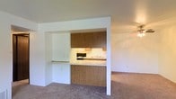 This single-floor residence offers a practical and straightforward layout designed to maximize usability. The living area is generously proportioned, featuring wall-to-wall carpeting that adds warmth and comfort underfoot. A ceiling fan overhead provides airflow and contributes to the room's clean, understated aesthetic.

The kitchen is framed by a distinctive built-in pass-through opening, which connects it visually to the living space and allows for easy interaction between the two areas. The kitchen is equipped with warm wood-toned cabinetry — both upper and lower — providing ample storage. A range with an overhead exhaust hood and a sink are neatly positioned within the kitchen's compact footprint. The countertop extends slightly into the pass-through area, functioning as a convenient breakfast bar or serving ledge.

The interior palette throughout is neutral and light, with white walls that help reflect light and create a bright, open atmosphere. The overall design is clean and uncluttered, making the space well-suited for personalization. The single-floor plan ensures easy accessibility and a seamless flow throughout the residence.