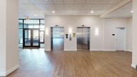 This thoughtfully designed lobby and common area spans a generous single floor, offering an open, gallery-like atmosphere that balances functionality with aesthetic appeal. The space is finished with warm wood-tone flooring throughout, crisp white walls, and a suspended drop ceiling with recessed lighting that creates a bright, welcoming ambiance.

Upon entry through floor-to-ceiling glass and dark-framed double doors, visitors are greeted by a spacious open floor plan that flows seamlessly across multiple zones. A central structural column adds architectural interest while naturally dividing the layout. Two stainless steel elevators are prominently positioned near the entrance, framed by wall sconces that lend a polished, refined quality.

Art is a defining feature of this space, with large-scale, colorful paintings and textured mixed-media works displayed prominently along the white walls, giving the interior a curated gallery feel. A cozy seating area is furnished with upholstered wingback chairs in soft grey arranged around small round side tables, anchored by a vibrant mosaic-style artwork in teal and gold. Elsewhere, a sculptural circular sofa in deep navy adds a bold, contemporary focal point.

Natural light filters in generously through the glass entryway, enhancing the clean, modern aesthetic throughout. The overall ambiance is polished, inviting, and sophisticated.