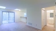 This single-floor residence offers a clean, open layout designed for comfortable everyday living. The main living area is bathed in natural light, courtesy of a ceiling skylight and multiple windows that brighten the neutral-toned walls and plush wall-to-wall carpeting beneath. A built-in pass-through counter connects the kitchen to the living space, creating a seamless flow between the two areas and making the layout feel both functional and social. The kitchen benefits from its own window, allowing additional natural light to filter through. A floor-mounted HVAC vent ensures efficient climate control throughout the living space. The interior is finished with clean white walls, white trim, and white baseboards, lending the space a crisp and well-maintained appearance. A louvered bi-fold closet door is visible along one wall, offering practical storage solutions while complementing the home's tidy aesthetic. The overall ambiance is light, airy, and welcoming, with a straightforward design that provides a versatile canvas suitable for a variety of furniture arrangements and personal styles.