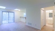 This single-floor residence offers a clean, open layout designed for comfortable everyday living. The main living area is bathed in natural light, courtesy of a ceiling skylight and multiple windows that brighten the neutral-toned walls and plush wall-to-wall carpeting beneath. A built-in pass-through counter connects the kitchen to the living space, creating a seamless flow between the two areas and making the layout feel both functional and social. The kitchen benefits from its own window, allowing additional natural light to filter through. A floor-mounted HVAC vent ensures efficient climate control throughout the living space. The interior is finished with clean white walls, white trim, and white baseboards, lending the space a crisp and well-maintained appearance. A louvered bi-fold closet door is visible along one wall, offering practical storage solutions while complementing the home's tidy aesthetic. The overall ambiance is light, airy, and welcoming, with a straightforward design that provides a versatile canvas suitable for a variety of furniture arrangements and personal styles.