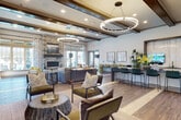 This well-appointed single-floor amenity space is designed to foster community and comfort through a thoughtfully curated blend of modern and transitional design elements. The expansive main lounge serves as the heart of the facility, anchored by a dramatic floor-to-ceiling stacked stone fireplace with a mounted television above. Exposed dark wood ceiling beams and striking circular pendant chandeliers add warmth and architectural character overhead, while wood-look plank flooring flows throughout the entire space. Large windows and glass doors flood the interior with natural light and offer views of an outdoor pool area, seamlessly connecting the interior to the exterior. Multiple seating vignettes — including plush sofas, upholstered accent chairs, and bar-height communal tables — provide versatile gathering options. A dedicated kitchenette features white cabinetry, dark countertops, open shelving, a beverage refrigerator, and coffee station, making it ideal for social events. A secondary lounge and game room area showcases another stacked stone accent wall with a wall-mounted television, plaid upholstered seating, and a shuffleboard table. A compact co-working nook with a high-top desk and curated artwork rounds out the space, offering a quiet spot for focused work within this vibrant communal environment.