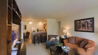 This single-story residence showcases an open-concept floor plan that integrates the living, dining, and kitchen areas into one cohesive space. The living area is comfortably appointed with a plush sofa in warm earth tones, a stylish blue accent chair with decorative pillows, and a dark wood coffee table adorned with an orchid centerpiece, contributing to an inviting ambiance. Recessed lighting and a ceiling fan provide both illumination and comfort throughout the space. The kitchen features dark wood cabinetry and is separated from the living area by a raised breakfast bar with dark counter stools, creating a natural division while maintaining visual openness. A white refrigerator and countertop appliances are visible, indicating a fully equipped kitchen. Adjacent to the main living space, a dining area with dark chairs is partially visible, offering a dedicated space for meals. The neutral-toned carpet and warm beige walls throughout create a cohesive, calming palette. Decorative wall art and carefully selected furnishings add character and a polished, lived-in feel to the overall space.