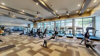 This generously sized fitness center occupies a single level and offers a comprehensive workout environment designed to accommodate a wide range of exercise needs. The space is defined by its expansive floor-to-ceiling windows along one side, flooding the interior with abundant natural light and providing open views of the surrounding outdoor areas, including landscaping and a neighboring pool area.

The ceiling features distinctive exposed dark beams that contrast with the warm neutral tones of the walls, creating a modern yet inviting aesthetic. Multiple ceiling fans are strategically positioned throughout, complementing the recessed lighting to maintain a comfortable atmosphere. Patterned, multi-tone carpet tiles in shades of tan, brown, and gray cover the floor throughout the entire space.

The fitness center is organized into clearly defined exercise zones. One section houses a robust selection of strength training machines, free weights, and a power rack, accompanied by wall-mounted televisions and mirrored walls. The cardio zone features treadmills, rowing machines, stationary bikes, ellipticals, and stair climbers positioned to take full advantage of the outdoor views. Built-in shelving near the entrance provides storage, and the overall layout ensures ample circulation space between equipment stations.