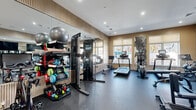 Located at 15 Washington Ave in Suffern, NY, this fitness center offers a thoughtfully designed exercise space that balances functionality with contemporary aesthetics. The room features warm, neutral-toned walls accented by vertical wood slat paneling, recessed lighting, and dark rubberized flooring that provides both durability and a sleek, professional appearance. Large windows line the perimeter, allowing ample natural light to fill the space while providing outdoor views that enhance the workout environment. The cardio zone is well-appointed with multiple treadmills, an elliptical trainer, a stair climber, and a stationary bike, each equipped with individual monitors. A dedicated strength training area includes a cable machine, adjustable weight benches, and a full rack of dumbbells. An organized equipment storage rack holds stability balls, foam rollers, and kettlebells, catering to functional fitness routines. Floor-to-ceiling mirrors are strategically placed to amplify the sense of space and assist with form. A wall-mounted flat-screen television adds convenience. Glass double doors open to an adjacent area, contributing to an open and connected feel. The overall ambiance is clean, modern, and motivating.