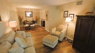 This single-floor space exudes a warm, welcoming ambiance anchored by rich hardwood flooring that flows seamlessly throughout the open-concept layout. The living area is thoughtfully appointed with plush, neutral-toned seating, including a comfortable sofa and an armchair paired with a matching ottoman, creating an ideal spot for relaxation. A classic floor lamp provides soft, ambient lighting that complements the warm beige tones of the walls. The living space opens naturally into the dining area, which features an elegant wrought-iron dining set beneath a decorative pendant light fixture. A mirror above the dining area adds depth and reflects light throughout the space, enhancing the sense of openness. Decorative touches such as dried botanical arrangements and framed wall art contribute to the refined yet approachable aesthetic. A small transitional area connects to additional functional spaces, including what appears to be a utility or storage area. Throughout, the consistent use of warm tones, quality flooring, and carefully selected furnishings create a cohesive and inviting atmosphere that balances everyday comfort with tasteful design.