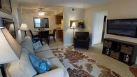 This single-floor residence offers a welcoming and functional layout with a seamlessly connected living, dining, and kitchen area. The living room is the heart of the space, featuring plush carpeted flooring, a comfortable sofa accented with decorative throw pillows, and a cushioned armchair positioned beside a classic floor lamp. A boldly patterned area rug anchors the seating arrangement and adds a vibrant touch to the neutral color palette of the walls and flooring. A ceiling fan with an integrated light fixture provides both comfort and illumination overhead. Just beyond the living area, the dining space accommodates a dark-toned table and chair set, complemented by a large framed mirror on the wall that reflects natural light and enhances the sense of openness. The kitchen, visible in the background, features warm wood-toned cabinetry and a raised breakfast bar, offering a convenient casual dining option. A hallway leading off the main living space suggests additional rooms beyond, contributing to the overall functional flow of the floor plan. The space maintains a warm, inviting ambiance with consistent neutral tones, soft carpeting, and thoughtful details throughout.