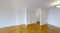 This single-floor space presents a clean, minimalist aesthetic defined by its crisp white walls and warm, honey-toned parquet hardwood flooring arranged in a classic geometric pattern throughout. The open layout creates a sense of spaciousness and flexibility, allowing for a variety of furnishing arrangements to suit different needs and lifestyles.

A set of built-in double closet doors provides convenient, integrated storage within the main living area, contributing to the uncluttered feel of the space. A hallway extends beyond the main room, leading to additional areas of the unit and featuring a ceiling-mounted light fixture that ensures consistent illumination throughout. The architectural lines are straightforward and understated, with smooth white ceilings and clean wall transitions that lend a timeless, neutral backdrop.

The overall ambiance is light, airy, and move-in ready, with the polished parquet floors adding warmth and character to the otherwise neutral palette. The simplicity of the design makes this space highly adaptable, offering a blank canvas well-suited for a range of interior styles and personal touches.