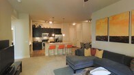This single-floor residence showcases a thoughtfully designed open-concept layout that seamlessly connects the kitchen, dining, and living areas into one cohesive and functional space. The kitchen is outfitted with dark cabinetry, stainless steel appliances — including a refrigerator, microwave, and range — and a granite-topped island with pendant lighting overhead. Vibrant orange bar stools line the island, adding a bold pop of color against the rich, dark cabinetry and neutral-toned walls. A tile backsplash and recessed track lighting further enhance the kitchen's modern aesthetic. Adjacent to the kitchen, the dining area features a sleek table and chairs, illuminated by additional track lighting. The living area is anchored by a large sectional sofa in a cool gray tone, accented with yellow and red throw pillows that complement the colorful artwork on display. A soft area rug defines the seating space, while the open floor plan creates an airy and inviting atmosphere throughout. Light-toned flooring and neutral walls provide a clean, versatile backdrop. The overall ambiance is contemporary and welcoming, blending functional design with carefully chosen décor accents.