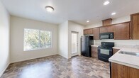 This efficiently designed one-bedroom, one-bathroom unit makes excellent use of its compact footprint across a single floor. The open living and dining area is bright and welcoming, featuring stone-look tile flooring that extends throughout the main living spaces and a large window that frames lush outdoor greenery, filling the room with natural light. A dedicated entry door provides direct access to the living area, which offers a clean, neutral palette with crisp white walls and flush ceiling lighting.

The kitchen is thoughtfully appointed with warm-toned medium brown cabinetry offering ample upper and lower storage, light countertops, and a full suite of black appliances including a refrigerator, electric range, and built-in microwave. Recessed lighting overhead ensures excellent task visibility, while a window near the entry keeps the space feeling open.

The bedroom features plush carpeting, neutral walls, and an open closet with a rod and shelf, providing practical storage. The bathroom continues the stone-look tile flooring from the main living area and includes a vanity with warm wood cabinetry, a single sink with light countertop, a combined tub and shower, and a wall-mounted heater. The consistent finishes throughout the unit create a cohesive and functional living environment.