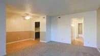 This single-floor residence offers a practical and comfortable layout designed for everyday living. The main living area is spacious and open, featuring soft neutral carpet and a ceiling fan that adds both function and a casual aesthetic to the room. A textured ceiling adds subtle character overhead, while large wall vents ensure efficient climate control throughout the space.

A half-wall partition connects the living area to the kitchen, which is outfitted with dark-toned cabinetry that provides a striking contrast against the lighter walls. The kitchen area transitions to hardwood-style flooring, adding a warm and durable surface underfoot. A two-tone wall treatment in the kitchen nook, featuring a tan lower half and a lighter upper half, adds a touch of classic design detail.

A hallway branching off from the main living space leads to additional areas of the home, with a wall-mounted light fixture providing a welcoming glow. The overall color palette throughout the unit is light and neutral, creating a clean and airy atmosphere. The layout flows naturally between spaces, making the home feel cohesive and easy to navigate. This residence offers a solid foundation for comfortable, functional everyday living.