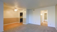 This single-floor residence offers a practical and comfortable layout designed for everyday living. The main living area is spacious and open, featuring soft neutral carpet and a ceiling fan that adds both function and a casual aesthetic to the room. A textured ceiling adds subtle character overhead, while large wall vents ensure efficient climate control throughout the space.

A half-wall partition connects the living area to the kitchen, which is outfitted with dark-toned cabinetry that provides a striking contrast against the lighter walls. The kitchen area transitions to hardwood-style flooring, adding a warm and durable surface underfoot. A two-tone wall treatment in the kitchen nook, featuring a tan lower half and a lighter upper half, adds a touch of classic design detail.

A hallway branching off from the main living space leads to additional areas of the home, with a wall-mounted light fixture providing a welcoming glow. The overall color palette throughout the unit is light and neutral, creating a clean and airy atmosphere. The layout flows naturally between spaces, making the home feel cohesive and easy to navigate. This residence offers a solid foundation for comfortable, functional everyday living.