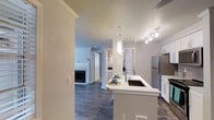This single-story residence features a well-appointed open-concept design that connects the kitchen and living areas seamlessly. The kitchen is a standout feature, showcasing a peninsula with a white quartz countertop, an undermount sink with a modern pull-down faucet, and pendant lighting overhead. Stainless steel appliances, including a full-size refrigerator, complement the white cabinetry, while track lighting provides ample illumination throughout. Dark hardwood-style flooring runs throughout the main living areas, creating a rich contrast against the light-toned walls and trim. Crown molding adds a refined architectural detail along the ceilings, which feature a textured finish. The living area is visible from the kitchen and includes a fireplace, serving as a focal point of the space. A hallway leads toward the bedroom and bathroom, offering a functional layout that maximizes the use of space. The overall ambiance is clean, modern, and welcoming, with neutral tones and contemporary fixtures creating a cohesive, move-in-ready aesthetic throughout the home.