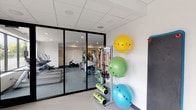 This single-floor fitness and yoga studio is thoughtfully designed to accommodate a range of exercise modalities in a clean, modern environment. The space is characterized by bright white drop-panel ceilings with recessed and flush-mounted lighting that creates an evenly lit, energizing atmosphere throughout.

A standout feature is the glass-partitioned workout zone, framed with sleek black-trim floor-to-ceiling glass panels that provide visual openness while defining separate training areas. This section houses cardio machines, including treadmills, and a variety of resistance training equipment. A dedicated accessories station lines one wall, featuring a multi-tiered rack stocked with stability balls in vibrant yellow, green, and blue, alongside foam rollers and resistance bands, keeping all essentials organized and within easy reach.

Adjacent to the main gym floor is a dedicated spin and cycling room, where a stationary bike takes center stage beneath a wall-mounted flat-screen television — ideal for guided cycling sessions or on-demand fitness content. The room features a calming gradient accent wall that transitions from white to teal, adding a refreshing visual element that complements the studio's wellness-focused atmosphere.

The overall design is clean and functional, with light-toned flooring, neutral walls, and a streamlined layout that maximizes usable space while maintaining an open, motivating environment suited for both individual training and group fitness activities.