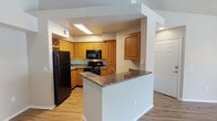 This single-floor residence offers a thoughtfully designed layout centered around a functional and inviting kitchen. The kitchen is a standout feature, showcasing warm honey-toned wood cabinetry that extends to the ceiling, providing generous storage throughout. A granite-topped peninsula serves as both a prep area and a casual dining or gathering space, elegantly separating the kitchen from the adjacent living area while maintaining an open, connected feel. The kitchen is equipped with black appliances, including a refrigerator, range, and built-in microwave, all complemented by the rich tones of the granite countertops. A decorative wine rack adds a charming detail to the upper cabinetry. The flooring throughout features light hardwood-style planks that lend warmth and continuity across the space. Recessed and flush-mounted lighting ensures the kitchen remains bright and well-lit. The open sightlines between rooms create a sense of spaciousness, making the layout ideal for both everyday living and entertaining. A closet with bi-fold doors is visible from the kitchen, hinting at practical storage solutions nearby. The overall ambiance is clean, warm, and move-in ready.