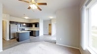 This single-floor space showcases a thoughtfully designed open-concept layout that seamlessly connects the living and kitchen areas. The living space features plush carpeting and a ceiling fan with integrated lighting, creating a comfortable and well-ventilated environment. The transition to the kitchen is marked by a shift to smooth hardwood-style flooring, lending a clean and modern aesthetic throughout.

The kitchen is outfitted with dark espresso cabinetry that provides a striking contrast against the neutral gray walls. Stainless steel appliances, including a range and microwave, are integrated alongside a kitchen island that offers additional prep space and storage. A deep basin sink is positioned beneath pendant lighting, adding a contemporary touch to the culinary space.

The neutral gray walls and light-toned ceilings create a cohesive, understated palette that allows natural light to brighten the interior. Doorways visible from the main living area lead to additional rooms, including what appears to be a bedroom with a walk-in closet, suggesting a well-organized and functional floor plan. White trim and clean baseboards throughout add a polished, finished look to the overall space.