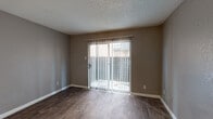 This single-story unit offers a functional and straightforward layout with distinct living, sleeping, kitchen, and bathroom areas. The living room serves as the central gathering space, finished with wood-look hardwood-style flooring and neutral taupe-toned walls. A full-length sliding glass door, dressed with vertical blinds, fills the room with natural light and provides direct access to an outdoor area. The front entry door opens into this same space, creating a welcoming and open feel.

The kitchen is positioned adjacent to the living area and features white cabinetry, speckled grey laminate countertops, and black appliances including a coil-burner electric range, over-the-range microwave, and refrigerator. A pass-through counter connects the kitchen to the main living space. The flooring in the kitchen continues the wood-look style seen throughout the common areas.

The bedroom is carpeted and benefits from two windows that allow natural light in from multiple directions. A ceiling fan with a light fixture is centrally mounted, providing both airflow and ambient lighting.

The bathroom includes a white vanity with a speckled countertop, a tub-shower combination with white tile surround and a curved curtain rod, and wood-look flooring consistent with the rest of the unit. Textured ceilings are present throughout the space.