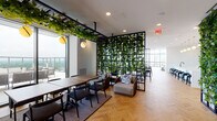 This upscale amenity floor is a masterclass in contemporary design, offering a seamless blend of social, recreational, and relaxation spaces. Throughout, warm herringbone-patterned hardwood flooring ties the various zones together, while expansive floor-to-ceiling glass curtain walls flood the interior with natural light and frame sweeping panoramic views of the surrounding cityscape and skyline.

The lounge area is anchored by a striking built-in fireplace framed in dramatic marble-veined stone, flanked by custom shelving and a large flat-screen display. Plush, neutral sofas with colorful accent pillows invite relaxed conversation. Nearby, a seating cluster of rich brown leather armchairs is positioned directly before the glass walls, maximizing the elevated views.

A dedicated game room features a full-size billiards table beneath soaring double-height ceilings supported by elegant cylindrical columns, creating an airy, open atmosphere. An intimate built-in banquette adds a cozy counterpoint to the grand scale.

The bar and kitchen area is centered around a long, white waterfall-edge island lined with black upholstered stools, complemented by open industrial shelving, a herringbone-tiled backsplash, and a cascade of warm globe pendant lights with brass accents. A separate lounge zone is thoughtfully divided by a lush living green wall on a black metal grid frame, adding a vibrant, organic element to the modern aesthetic. An outdoor terrace extends the entertaining space beyond the glass.