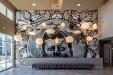 This single-floor space makes an immediate impression with its dramatic double-height entrance lobby, where floor-to-ceiling glass doors and expansive clerestory windows flood the interior with natural light. A large-scale, mixed-media mural dominates one wall of the entrance area, blending intricate black-and-white illustration with photorealistic imagery and extending from floor to ceiling, serving as a defining artistic focal point. Clusters of spherical globe pendant lights hang at varying heights throughout, complementing the warm yellow ceiling accents and adding a sculptural quality to the space.

The lounge area transitions seamlessly from the entry, featuring a bold red sectional sofa, organic wood coffee tables, and patterned area rugs over large-format tile flooring. A textured stone fireplace column anchors the interior lounge, with a wall-mounted television above and a warm wood beam with integrated lighting overhead. Contemporary white egg-style lounge chairs and a red bench complete the seating arrangement, lending a modern, curated aesthetic.

Adjacent to the lounge sits a café station with bar-height seating, dark wood cabinetry, and pendant lighting. Glass-partition walls and sliding doors define transitional zones within the space while maintaining visual openness throughout. The overall ambiance is sophisticated, welcoming, and design-forward.
