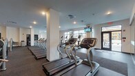 Located at 40 Hutchinson Ave in Columbus, Ohio, this expansive fitness center is thoughtfully designed to accommodate a full range of workout needs across a single floor. The space is organized into distinct training zones, each outfitted with professional-grade equipment and finished with durable rubber flooring suitable for high-intensity use.

The cardio zone anchors a large central area and features multiple rows of treadmills and elliptical machines, each equipped with individual screens for entertainment. Large windows line the perimeter, flooding the space with natural light and offering views of the surrounding area. Mounted televisions throughout ensure users stay engaged during their workouts.

A dedicated cycling studio offers a focused environment for spin sessions, complete with several stationary bikes, a wall-spanning mirror, and a mounted display screen. Adjacent areas house an extensive strength training section, lined with a comprehensive selection of cable machines, selectorized resistance equipment, and multi-use stations positioned along mirrored walls.

A free weights area provides a power rack, adjustable benches, and a full dumbbell rack. A functional training zone rounds out the facility, featuring TRX suspension trainers, medicine balls, and additional accessory equipment. The entrance corridor provides a clean, modern welcome with dark-framed glass doors and polished finishes throughout.