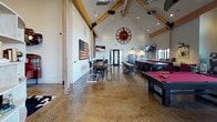 Located in Pullman, WA, this single-story amenity building is designed with a rustic-industrial aesthetic that blends exposed timber beam construction, polished concrete floors, and bold decorative elements throughout. The structure's architectural centerpiece is its soaring vaulted ceiling supported by heavy wood trusses with steel bracket hardware, lending the main gathering area a barn-inspired character with a modern edge.

The primary social hall is expansive and open, featuring large floor-to-ceiling windows along one wall that flood the interior with natural light and provide views of the surrounding neighborhood. This area is furnished with a pool table, foosball table, ping pong table, bar-height seating, and multiple flat-screen televisions, making it well-suited for entertainment and recreation. An American flag art piece and an oversized decorative wall clock contribute to the space's distinctive personality.

The reception and leasing area includes a reclaimed wood-paneled front desk beneath geometric pendant lighting, set against a bold red accent wall branded with "The Hills." Adjacent to this, glass-walled private offices provide transparency while maintaining a functional workspace.

A secondary room with polished concrete floors, barn-style industrial lighting, open shelving, and a mounted display offers additional flexible programming space. An entry foyer features stacked stone accent walls, patterned carpet tiles, and tall windows that introduce natural light from the exterior.