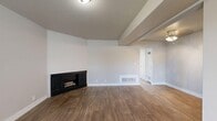 This efficient and thoughtfully designed single-floor unit offers a functional layout comprising a living room, bedroom, bathroom, and kitchen. The living area features wood-style plank flooring and a charming corner fireplace with a classic dark surround, creating a warm and inviting focal point. Natural light enters through a window fitted with blinds, accompanied by a wall-mounted HVAC unit for year-round comfort. The galley-style kitchen is well-equipped with white shaker cabinetry, quartz countertops, a stainless steel double-basin sink, a dishwasher, and black appliances including a refrigerator and range. A window above the sink brings in additional natural light. A hallway with wood-style flooring connects the main living areas to the bedroom and bathroom. The bathroom includes a white vanity with quartz countertops and brushed nickel fixtures. The bedroom is carpeted with soft, neutral-toned flooring and features a ceiling fan with light fixture, a closet, and a sliding glass door that opens to an outdoor view, allowing for natural light and ventilation. Neutral wall tones and clean white trim are consistent throughout, lending the space a cohesive and move-in-ready aesthetic.
