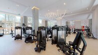 This expansive fitness center is thoughtfully designed to offer a comprehensive wellness experience across a generous, open single-floor layout. The space is organized into distinct functional zones, each tailored to specific training styles while maintaining a cohesive, upscale aesthetic throughout.

The main strength and cardio area features an impressive array of professional-grade machines, including treadmills, stationary bikes, rowing machines, and a full range of resistance equipment. Tall, floor-to-ceiling windows flood the space with natural light and offer views of the surrounding streetscape, while large wall mirrors amplify the sense of openness. A sculptural linear chandelier anchors the ceiling, adding an elegant design focal point above the workout floor.

A dedicated functional training zone offers suspension trainers, stability balls, medicine balls, and open floor space ideal for dynamic movement and group sessions. Warm-toned framed glass partitions define this area while preserving visual connectivity throughout. An additional free weights section houses a full dumbbell rack alongside a power rack and benches.

Connecting the zones is a tranquil entry corridor distinguished by a dramatic living green wall, where lush vertical panels of ferns and foliage introduce a biophilic element that softens the environment. Light-toned hardwood and tile flooring run throughout, complementing the clean white walls and exposed concrete columns that lend the space an architectural sophistication. Direct access to an outdoor courtyard further enhances the facility's connection to nature.