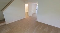 This two-floor space is characterized by its clean, neutral aesthetic and practical layout. The interior features wall-to-wall beige carpeting throughout the main living areas, providing a soft and cohesive foundation across the rooms. The walls are painted in a crisp white, contributing to a bright and open atmosphere.

Visible from the main carpeted room is an attached bathroom, accessible through a standard interior door. The bathroom is finished with tile flooring, a pedestal sink, and a mirror, offering a clean and functional design. A ceiling-mounted light fixture illuminates the bathroom space.

The adjoining room, partially visible through an open passage, continues the same carpeted flooring and white walls, maintaining a consistent aesthetic throughout. Natural light filters into this connected area through what appears to be a large window with vertical blinds, enhancing the overall brightness of the space.

A ceiling fan is present in the secondary room, adding to the functional features of the unit. The overall ambiance is straightforward and well-maintained, with a neutral palette that provides a versatile backdrop suitable for a variety of furnishing styles.
