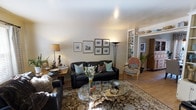 This two-storey space presents a thoughtfully designed interior with a welcoming living area as a central feature. The living room is furnished with a dark leather sofa accented by teal and patterned cushions, complemented by a ornate upholstered armchair that adds an eclectic touch. A glass-topped coffee table with a natural wood base serves as a focal point over a richly patterned area rug, grounding the seating arrangement.

The walls are adorned with a curated gallery of framed artwork, contributing to an cultivated, lived-in aesthetic. Built-in white shelving units flank a connecting hallway, offering both display and storage space for books and decorative objects. Warm lighting from floor and table lamps enhances the ambient glow of the room, creating a cozy atmosphere. Light hardwood flooring extends through the space, adding warmth underfoot.

Lush indoor plants introduce natural elements, while the eclectic mix of furnishings and accessories — including sculptural objects and ceramics — lends the space a well-traveled, personalized character. The layout flows naturally, suggesting well-considered spatial planning across both levels.