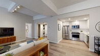 This single-floor residence offers a well-designed open-concept layout with distinct functional zones connected by light-toned hardwood flooring. The living area is generously proportioned, featuring notably tall ceilings that enhance the sense of volume, and is illuminated by multiple large windows fitted with horizontal blinds. A compact dining nook adjacent to the living space accommodates a small table and doubles as a workspace, maximizing the utility of the layout.

The kitchen is efficiently designed with white shaker-style cabinetry extending to the ceiling, white quartz countertops, a subway tile backsplash, and a full suite of stainless steel Whirlpool appliances including a refrigerator, range, and over-the-range microwave. Recessed lighting highlights the clean, bright aesthetic of the space.

A dedicated laundry closet houses a stacked washer and dryer unit, providing in-unit convenience. The bedroom is spacious with plush carpeting, soaring ceilings, direct access to a walk-in closet, and an en-suite bathroom. The bathroom features a frameless glass walk-in shower enclosed in crisp white subway tile with a built-in corner shelf. Neutral gray walls and consistent modern finishes unify the overall ambiance throughout.