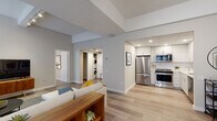 This single-floor residence offers a well-designed open-concept layout with distinct functional zones connected by light-toned hardwood flooring. The living area is generously proportioned, featuring notably tall ceilings that enhance the sense of volume, and is illuminated by multiple large windows fitted with horizontal blinds. A compact dining nook adjacent to the living space accommodates a small table and doubles as a workspace, maximizing the utility of the layout.

The kitchen is efficiently designed with white shaker-style cabinetry extending to the ceiling, white quartz countertops, a subway tile backsplash, and a full suite of stainless steel Whirlpool appliances including a refrigerator, range, and over-the-range microwave. Recessed lighting highlights the clean, bright aesthetic of the space.

A dedicated laundry closet houses a stacked washer and dryer unit, providing in-unit convenience. The bedroom is spacious with plush carpeting, soaring ceilings, direct access to a walk-in closet, and an en-suite bathroom. The bathroom features a frameless glass walk-in shower enclosed in crisp white subway tile with a built-in corner shelf. Neutral gray walls and consistent modern finishes unify the overall ambiance throughout.