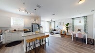 This single-floor model residence in Port Orange, Florida showcases a thoughtfully designed open-concept layout that seamlessly connects the kitchen, dining, and living areas. The kitchen is outfitted with white shaker-style cabinetry, granite countertops, stainless steel appliances including a refrigerator, range, microwave, and dishwasher, and a central island with bar seating — ideal for casual dining and entertaining. Wood-look flooring extends throughout the main living areas, adding warmth and visual continuity. The living room is bright and airy, benefiting from large windows and sliding glass door access, while a ceiling fan enhances comfort. The adjacent dining area features a pendant light fixture and ample space for a full dining set. The coastal-inspired décor, with teal and sage accents, patterned curtains, and curated artwork, reflects the relaxed Florida lifestyle. Two well-proportioned bedrooms offer plush carpeting, natural light from windows with blinds, and comfortable closet space. The bathroom includes wood-look flooring, a shower with curtain, and granite-topped vanity. Additional utility space is also present. Overall, the layout is functional and inviting, making it an excellent representation of modern residential living in Port Orange.
