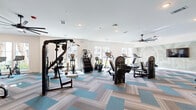 Located at 100 Wilton Cir S in Palm Coast, Florida, this fitness center offers a generously sized, single-floor workout space designed to accommodate a full range of exercise routines. The open layout is finished with stylish herringbone-patterned carpet tiles in neutral tones accented with aqua blue, lending a modern, polished aesthetic throughout.

The cardio zone, positioned along a wall of large windows, features multiple treadmills, ellipticals, and a stair climber, all bathed in abundant natural light with views of lush landscaping. Large ceiling fans are installed throughout, promoting air circulation and comfort during workouts.

The strength training area is equally well-appointed, housing a cable multi-gym, a squat and power rack, a dedicated dumbbell rack, adjustable benches, and a variety of selectorized resistance machines targeting multiple muscle groups. Full-length mirrors line one wall, enhancing the sense of space and allowing users to monitor their form.

A built-in open shelving unit near the back of the room provides organized storage for yoga blocks, foam rollers, and other fitness accessories. An adjacent room, accessible through a glass-paneled door, offers additional utility space. A decorative palm tree wall mural adds a Florida-inspired touch, completing the bright and inviting atmosphere of this comprehensive fitness facility.