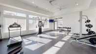 This spacious fitness center is designed with a clean, modern aesthetic, characterized by crisp white walls, soaring ceilings, and large windows dressed with horizontal blinds that flood the space with natural light. The open layout provides ample room for movement, while durable dark rubberized flooring offers both functionality and a polished, contemporary look throughout.

The strength-training area is well-appointed, featuring a multi-functional cable machine, a full rack with barbell and weight plates, an adjustable bench, and a tiered dumbbell rack stocked with a wide range of weights. Wall-mounted flat-screen televisions are positioned throughout the space, keeping users informed and entertained during workouts.

The cardio zone is equally comprehensive, housing multiple elliptical trainers and a treadmill, all positioned to take advantage of the natural light streaming in from the surrounding windows. Ceiling fans are installed overhead to maintain airflow and comfort during intense sessions. A glass-paneled door connects the fitness area to an adjacent lounge space, offering a comfortable area for rest and recovery. The overall ambiance is bright, airy, and motivating, making this a highly functional and welcoming fitness facility.