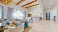 Located at 3514 Aspire Circle in Cape Coral, Florida, this beautifully appointed single-floor space showcases a coastal-contemporary design highlighted by soaring vaulted ceilings with exposed natural wood beams and light hardwood flooring throughout. The expansive open-concept main area seamlessly connects a generous lounge zone with multiple seating arrangements, a kitchen and bar area featuring a large island with marble-style countertops, a decorative tile backsplash, and pendant chandelier lighting. The lounge is further defined by elegant built-in shelving with dark cabinetry, a wall-mounted display, and a striking woven pendant light fixture that anchors the space. A cozy sitting nook with deep teal accent walls, floor-to-ceiling windows, and lush tropical outdoor views offers an inviting retreat. The space also includes a light-filled dining area with tall wraparound windows framing palm tree views, and a dedicated private office accessed through glass-paneled doors. A wide, well-lit corridor connects additional rooms with glass-door entries. The expansive covered outdoor terrace, accessible via sliding glass doors, features a ceiling fan, open-air lounge and dining areas, and panoramic views of the surrounding community and landscaping.