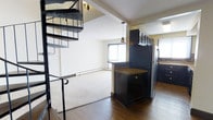 This two-floor residence offers a functional and visually engaging layout that combines open-concept living with practical design. The main level immediately draws attention with a bold spiral staircase constructed from dark metal, serving as both a functional element and an architectural focal point. Hardwood flooring anchors the main living and kitchen areas, transitioning seamlessly to plush carpet in the living room, adding warmth and comfort underfoot.

The kitchen is outfitted with dark cabinetry that extends to the upper wall, offering generous storage capacity. A central island with a granite-toned countertop provides additional prep space and acts as a natural divider between the kitchen and living areas. Stainless steel appliances complete the kitchen's sleek, modern aesthetic. Pendant lighting above the island adds focused illumination and a contemporary design touch.

The adjoining living area is bright and open, benefiting from natural light filtering through a large window. A baseboard heating system runs along the perimeter, ensuring comfortable temperatures throughout the seasons. Textured ceilings and light-toned walls contribute to a clean, neutral palette that complements the dark cabinetry and metalwork. The second floor, accessible via the spiral staircase, extends the living space further, making this a well-rounded multi-level residence.