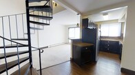 This two-floor residence offers a functional and visually engaging layout that combines open-concept living with practical design. The main level immediately draws attention with a bold spiral staircase constructed from dark metal, serving as both a functional element and an architectural focal point. Hardwood flooring anchors the main living and kitchen areas, transitioning seamlessly to plush carpet in the living room, adding warmth and comfort underfoot.

The kitchen is outfitted with dark cabinetry that extends to the upper wall, offering generous storage capacity. A central island with a granite-toned countertop provides additional prep space and acts as a natural divider between the kitchen and living areas. Stainless steel appliances complete the kitchen's sleek, modern aesthetic. Pendant lighting above the island adds focused illumination and a contemporary design touch.

The adjoining living area is bright and open, benefiting from natural light filtering through a large window. A baseboard heating system runs along the perimeter, ensuring comfortable temperatures throughout the seasons. Textured ceilings and light-toned walls contribute to a clean, neutral palette that complements the dark cabinetry and metalwork. The second floor, accessible via the spiral staircase, extends the living space further, making this a well-rounded multi-level residence.