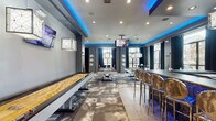 Located at 3400 S Bowman Rd in Little Rock, Arkansas, this single-floor entertainment lounge is designed as a sleek, modern gathering space with a distinctly upscale aesthetic. The interior is defined by a sophisticated gray color palette accented by striking blue LED cove lighting that runs along the ceiling perimeter, creating a vibrant and energetic ambiance throughout.

The space is anchored by a well-appointed bar area featuring a long counter with blue glass mosaic tile detailing, lined with polished chrome bar stools. A display shelf stocked with bottles and an embedded flat-screen TV complete the bar setup, making it a natural social focal point. Large floor-to-ceiling glass doors provide natural light and a view to adjacent areas.

The main gaming area is generously proportioned and equipped with multiple billiards tables and a full-length shuffleboard table, all finished in a cohesive modern style. Stylish pendant light fixtures hang above the game tables, while wall-mounted flat-screen televisions are positioned throughout for optimal viewing. Abstract patterned carpet tiles add visual texture and define the gaming zones, complemented by tile flooring along the bar corridor.

High-top tables and sleek bar-height seating are arranged throughout, offering flexible social configurations. Decorative spherical wall sculptures and dark drapery panels add a polished, lounge-like character to the overall design.