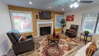 Located on Nautilus Circle in Fort Worth, TX, this single-story property offers a well-designed interior centered around a spacious, open-concept living area. The room is anchored by a classic tile-surround fireplace with a white-painted mantle, above which a wall-mounted flat-screen television is positioned as a focal point. Warm golden-yellow accent walls contrast with neutral white and gray tones throughout, contributing to an inviting and energetic atmosphere.

Wood-look plank flooring extends throughout the living space, providing a cohesive and durable foundation. Recessed lighting and ceiling fans ensure consistent illumination and airflow across the room. Multiple windows fitted with horizontal blinds allow natural light to filter in from multiple directions, brightening the interior noticeably.

A dedicated workspace is integrated seamlessly into the layout, featuring a built-in dark wood desk with granite-style countertop and cabinet storage. An arched passageway connects this area to the kitchen, which is visible in the background and features dark cabinetry, stainless steel appliances, pendant lighting, and a breakfast bar. The overall layout promotes a natural flow between living, working, and dining areas, making this a functional and visually cohesive single-floor space.