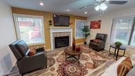 Located on Nautilus Circle in Fort Worth, TX, this single-story property offers a well-designed interior centered around a spacious, open-concept living area. The room is anchored by a classic tile-surround fireplace with a white-painted mantle, above which a wall-mounted flat-screen television is positioned as a focal point. Warm golden-yellow accent walls contrast with neutral white and gray tones throughout, contributing to an inviting and energetic atmosphere.

Wood-look plank flooring extends throughout the living space, providing a cohesive and durable foundation. Recessed lighting and ceiling fans ensure consistent illumination and airflow across the room. Multiple windows fitted with horizontal blinds allow natural light to filter in from multiple directions, brightening the interior noticeably.

A dedicated workspace is integrated seamlessly into the layout, featuring a built-in dark wood desk with granite-style countertop and cabinet storage. An arched passageway connects this area to the kitchen, which is visible in the background and features dark cabinetry, stainless steel appliances, pendant lighting, and a breakfast bar. The overall layout promotes a natural flow between living, working, and dining areas, making this a functional and visually cohesive single-floor space.