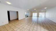 This single-floor space presents a generous, open-plan layout finished with light beige ceramic floor tiles laid in a classic grid pattern throughout, contributing to a clean and cohesive aesthetic. The main living area is notably spacious, offering an expansive feel that lends itself well to a variety of furniture arrangements and uses.

Natural light is a key feature, with multiple windows along the exterior wall, including a large sliding glass door fitted with vertical blinds and smaller barred windows providing additional ventilation and light. Together, these openings create a bright and airy atmosphere throughout the day.

The ceilings feature a textured popcorn finish and are equipped with flush-mounted light fixtures, ensuring consistent artificial lighting across the space. The walls are painted in a crisp white, further enhancing the sense of openness and light.

A doorway visible on the left side of the main area leads to an adjoining room or alcove, adding functional versatility to the layout. Overall, the space presents a straightforward, well-lit interior that is move-in ready and adaptable to various purposes.
