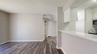 This single-floor, one-bedroom, one-bathroom unit offers a practical and well-organized layout across a modest footprint. The combined kitchen and living room serves as the heart of the space, featuring dark wood-style vinyl flooring throughout and neutral gray walls that create a clean, contemporary aesthetic. The living area is highlighted by a classic fireplace with a white mantel, vertical blind-covered sliding glass doors that allow natural light to filter in, and track lighting overhead. The kitchen is efficiently laid out in a galley style, equipped with white cabinetry, a stainless steel refrigerator, and a stove, neatly separated from the living area by a partial wall with an overhead cabinet pass-through. The bedroom offers plush carpeting, a ceiling fan with integrated lighting, and a window with blinds for natural light. A mirrored door leads to a dedicated walk-in closet fitted with multi-level shelving rods, providing generous storage. The bathroom includes a full tiled tub-shower combination, a toilet, and a vanity. Textured popcorn ceilings are present throughout the unit. A dedicated closet area adds additional storage convenience.